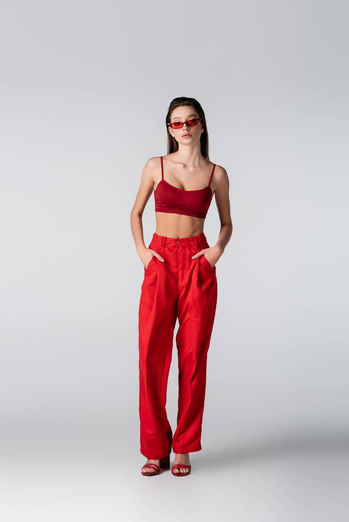 longueur totale de jeune modèle en lunettes de soleil et tenue rouge posant avec les mains dans les poches sur gris - Photo, image