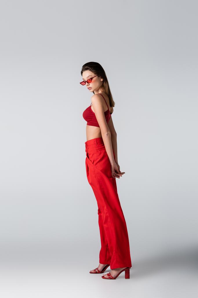 πλήρες μήκος της μελαχρινής γυναίκας με γυαλιά ηλίου και κόκκινο ντύσιμο ποζάρουν σε γκρι - Φωτογραφία, εικόνα