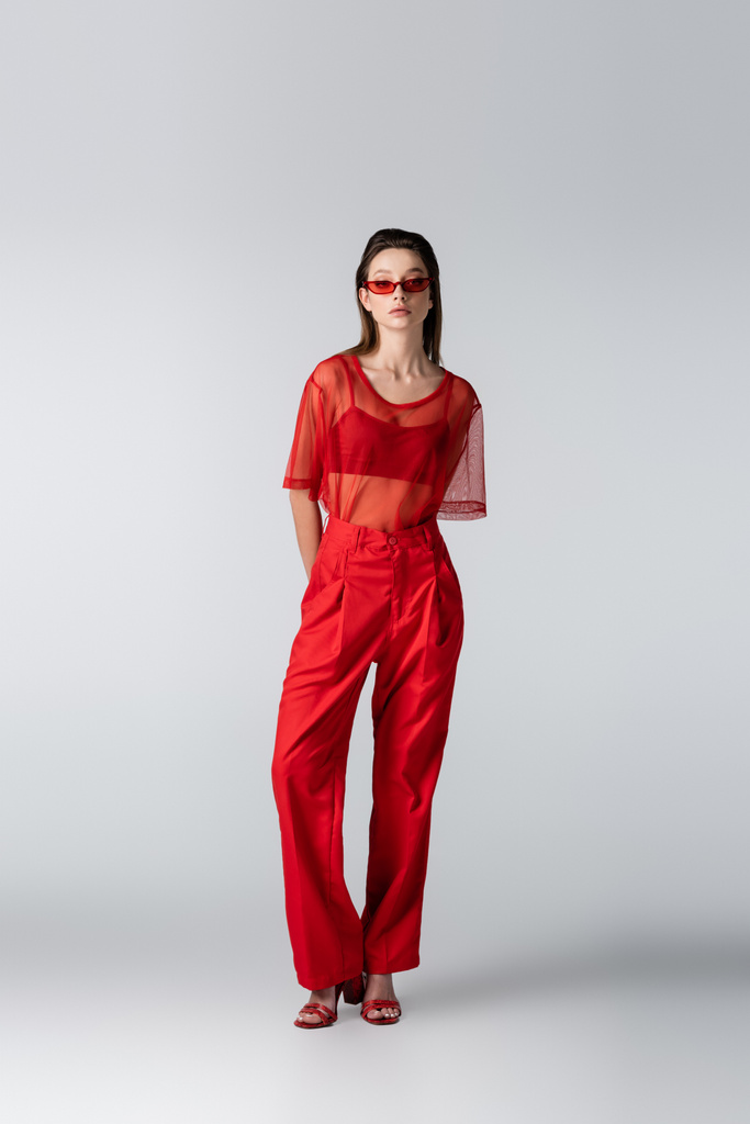 longitud completa de modelo joven en gafas de sol y traje rojo de moda posando en gris - Foto, Imagen
