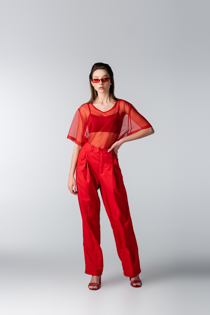 pełna długość młodej modelki w czerwonym modnym stroju i okularach przeciwsłonecznych pozujących z dłonią na biodrze na szarości - Zdjęcie, obraz