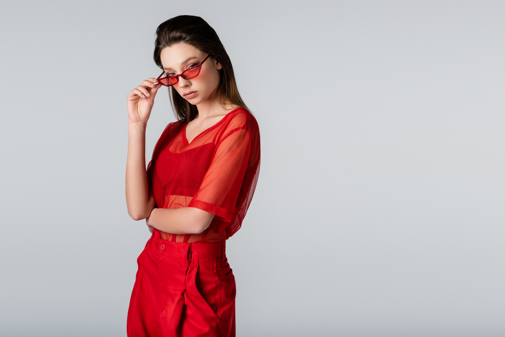 giovane donna in abito rosso alla moda che regola gli occhiali da sole mentre posa isolata sul grigio - Foto, immagini