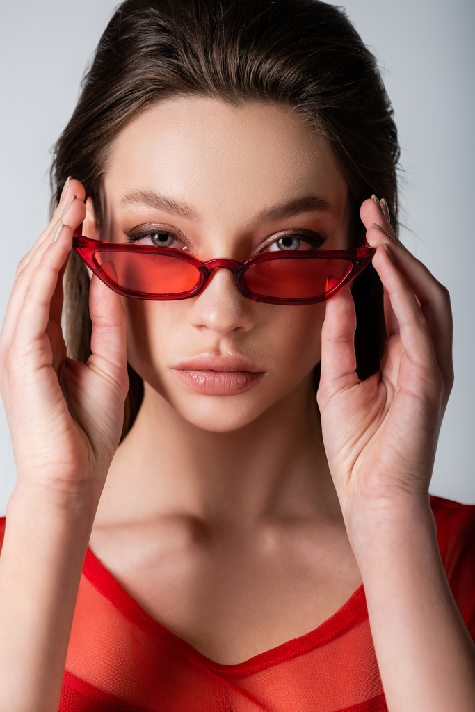 young model adjusting red trendy sunglasses isolated on grey - Photo, Image