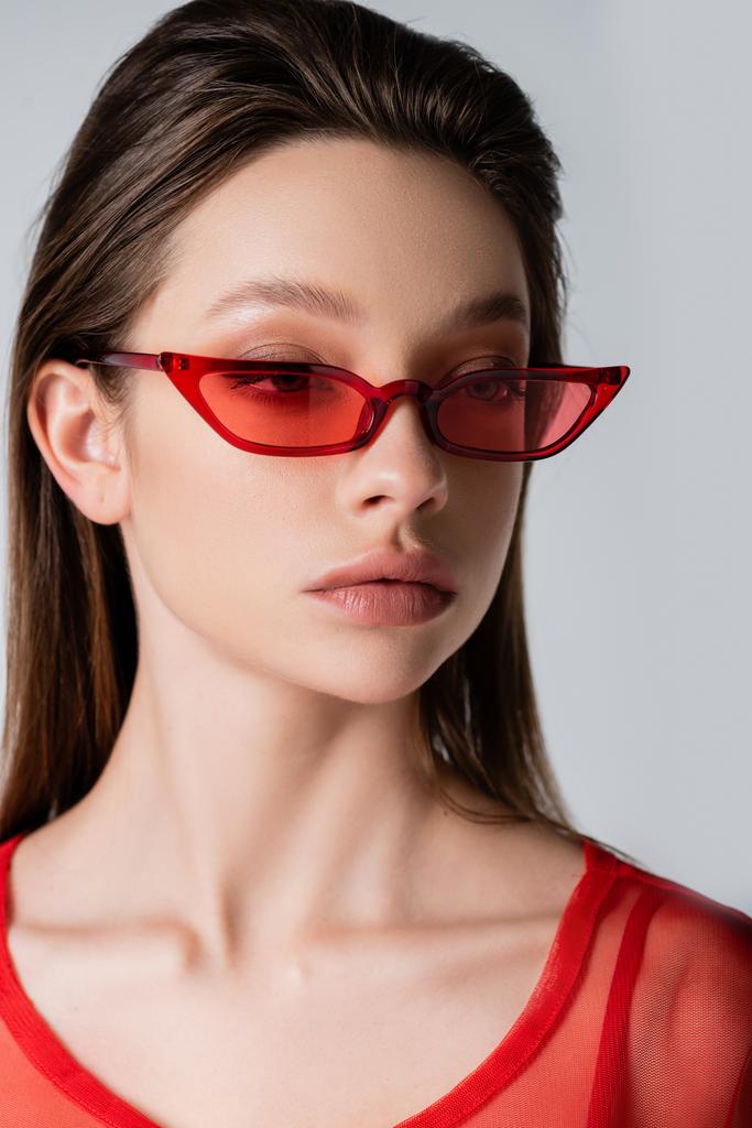 молодая модель в красных модных солнцезащитных очках, изолированных на сером - Фото, изображение