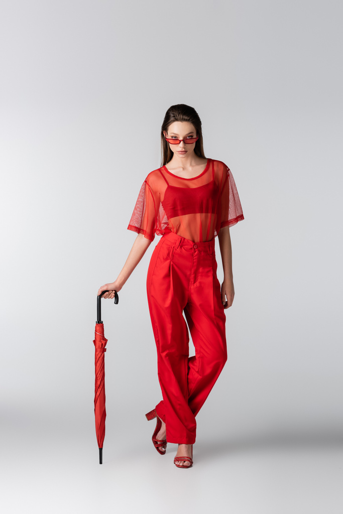 πλήρες μήκος του νεανικού μοντέλου σε κόκκινο μοντέρνο ντύσιμο και γυαλιά ηλίου ποζάρουν με ομπρέλα σε γκρι - Φωτογραφία, εικόνα