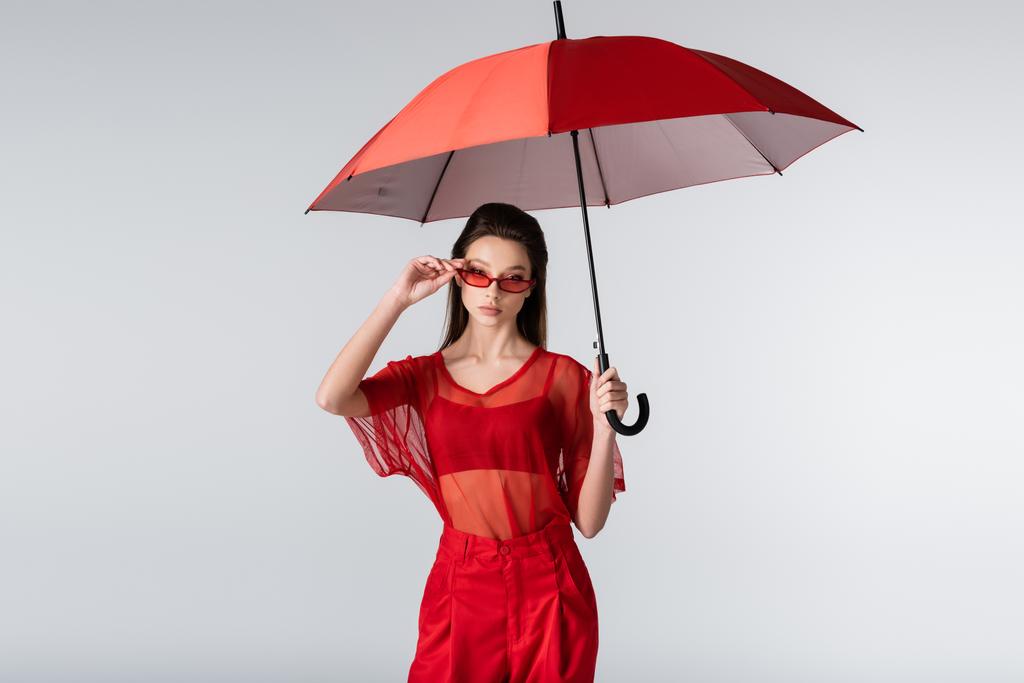 mujer joven en traje de moda ajustando gafas de sol rojas y de pie bajo paraguas aislado en gris - Foto, imagen