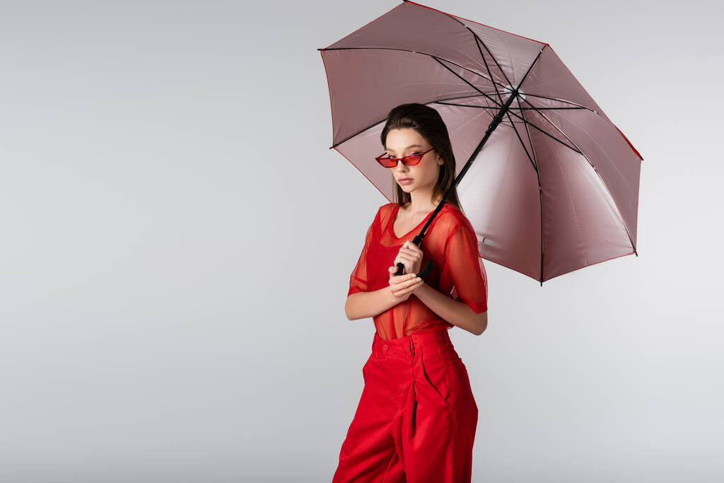 молодая женщина в красном костюме и солнцезащитных очках, стоящая под зонтиком изолированная на сером - Фото, изображение