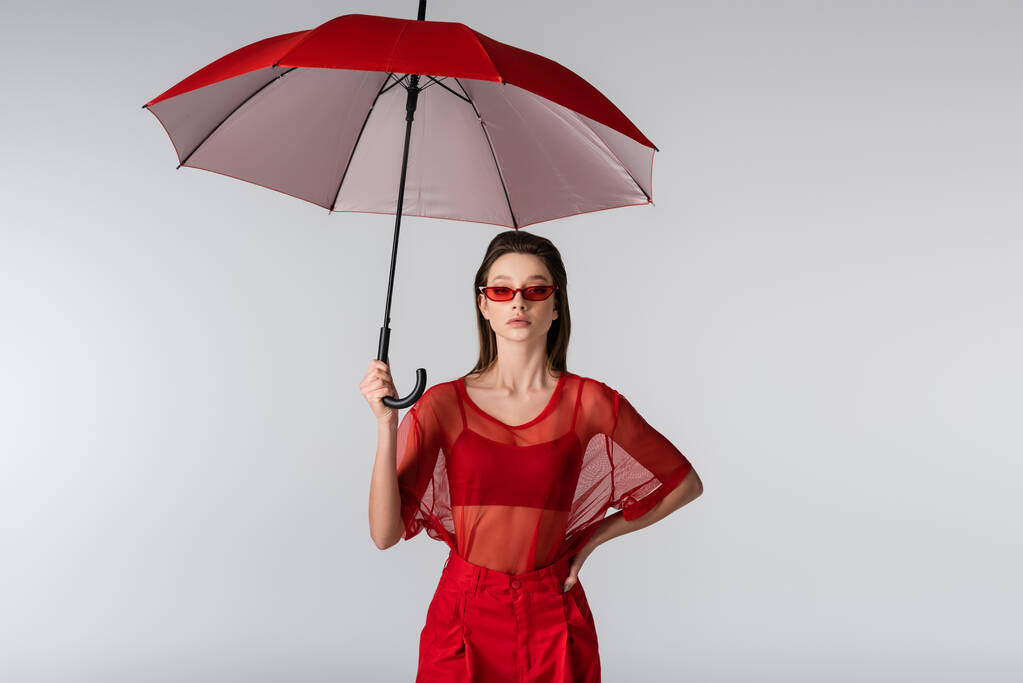 young woman in trendy outfit and red sunglasses standing with hand on hip under umbrella isolated on grey - Photo, Image