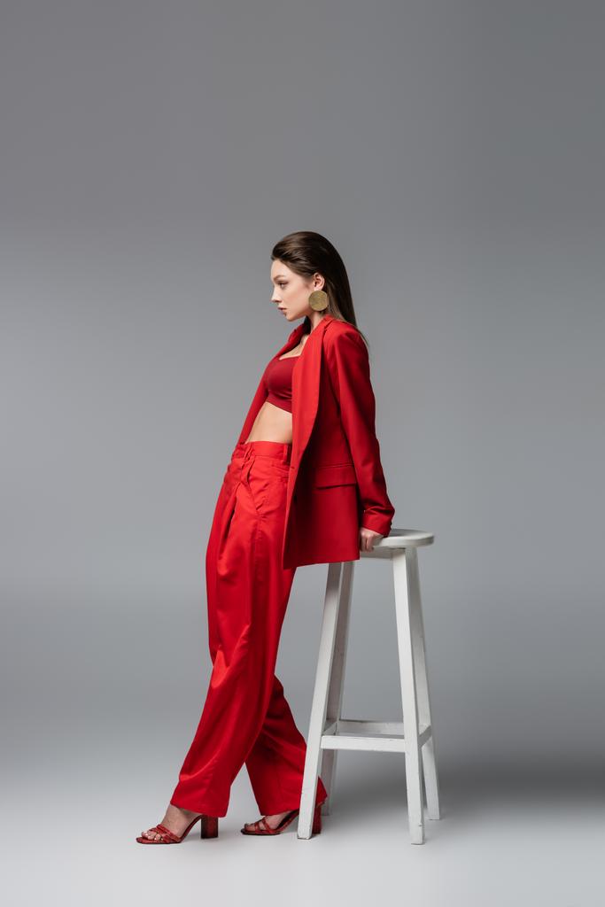 volledige lengte van jonge vrouw in trendy rood pak staan in de buurt van witte stoel op donkergrijs - Foto, afbeelding
