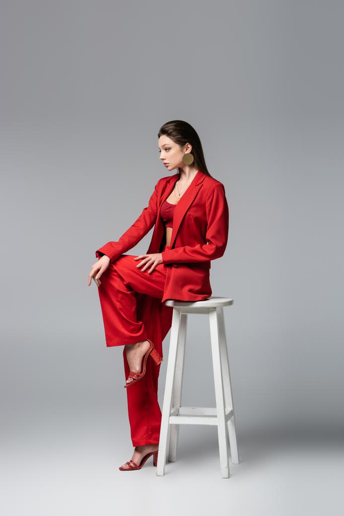 トレンディな赤いスーツを着た若い女性の完全な長さ黒灰色の上に白い椅子に座って - 写真・画像