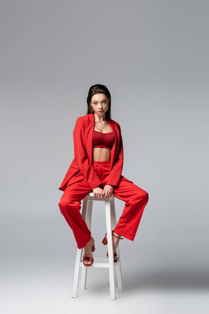 full length of young woman in red suit and shoes sitting on white chair on dark grey - Photo, Image