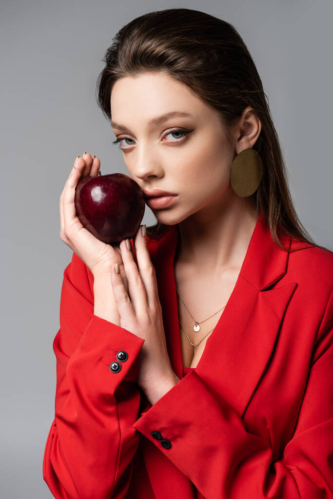 молода жінка в червоному блістері і сережці тримає яблуко ізольовано на сірому
  - Фото, зображення