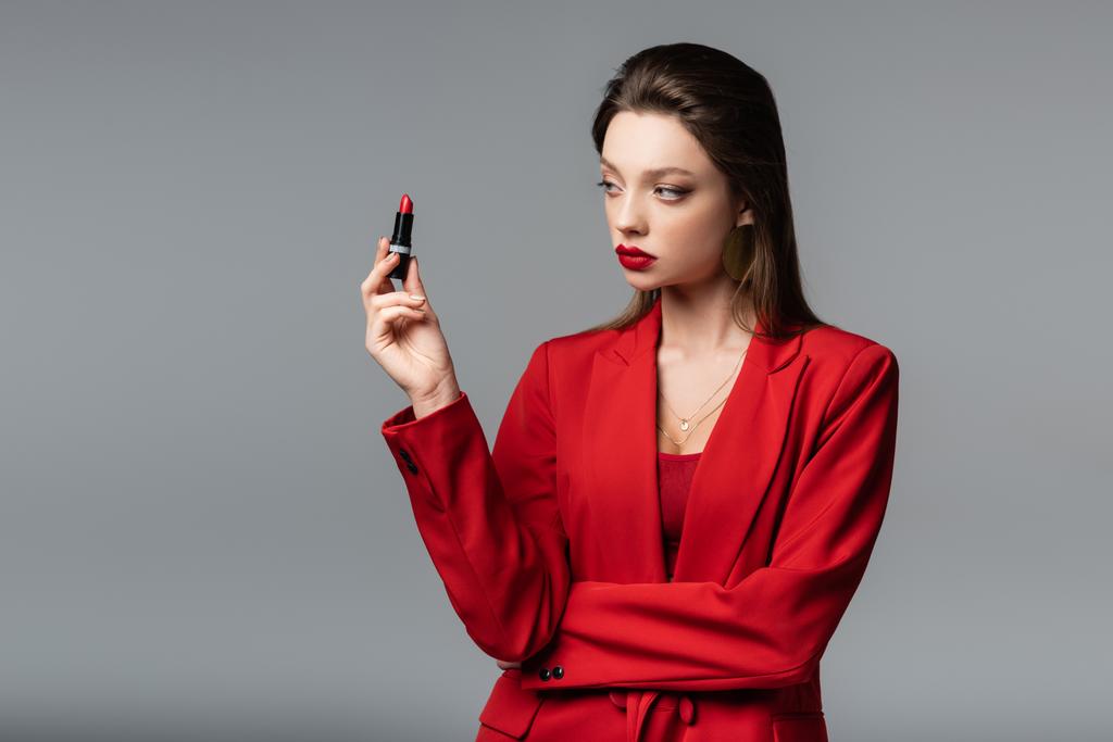junge Frau im roten Blazer blickt auf Lippenstift vereinzelt auf grau - Foto, Bild