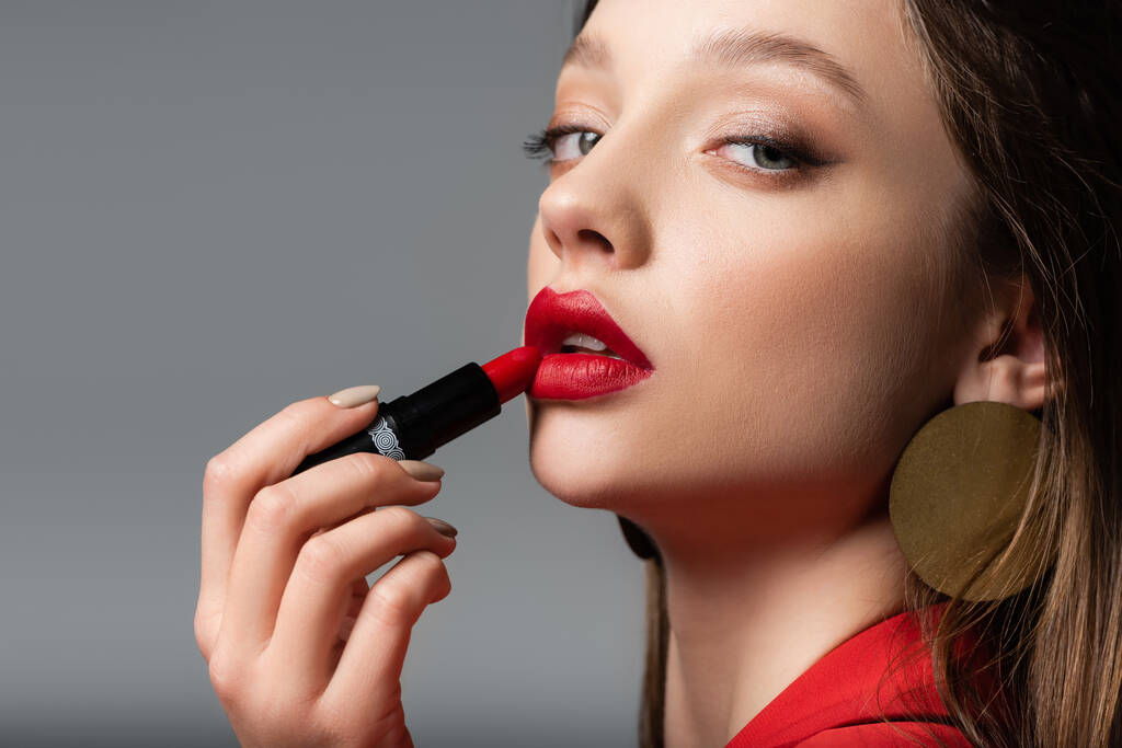 Nahaufnahme junger Frau, die roten Lippenstift isoliert auf grau aufträgt - Foto, Bild