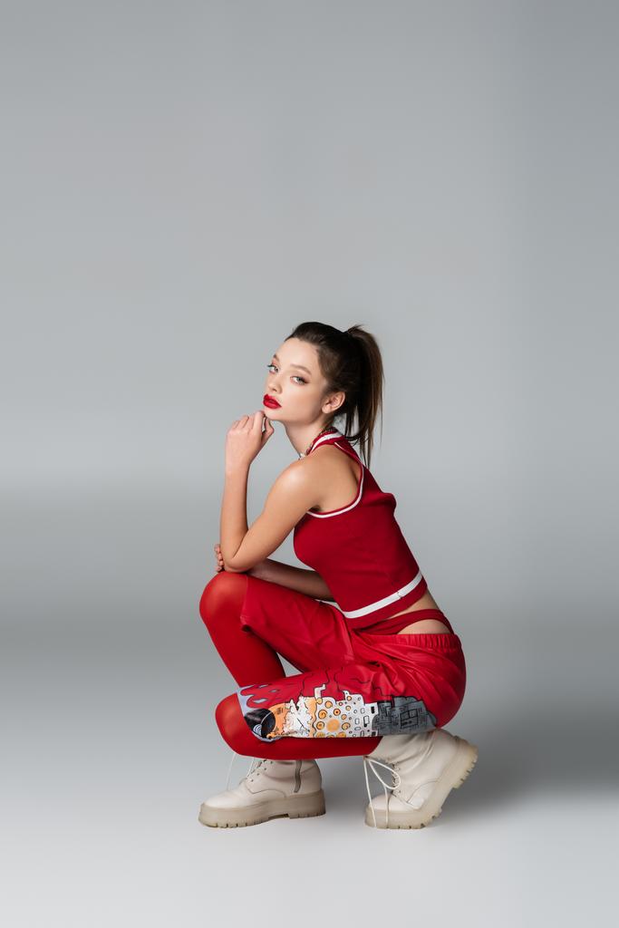 pełna długość stylowa młoda kobieta w czerwonym stroju sportowym i buty siedzi podczas pozowania na szarym - Zdjęcie, obraz