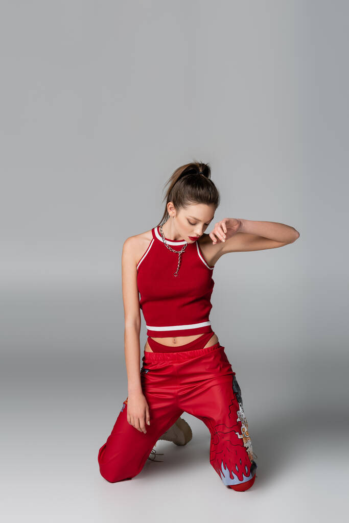 jovem elegante em roupas esportivas vermelhas em pé sobre os joelhos em cinza - Foto, Imagem