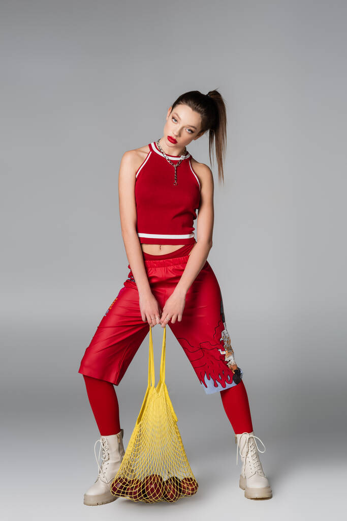 comprimento total da jovem mulher em roupa esportiva vermelha segurando saco de cordas com maçãs em cinza - Foto, Imagem
