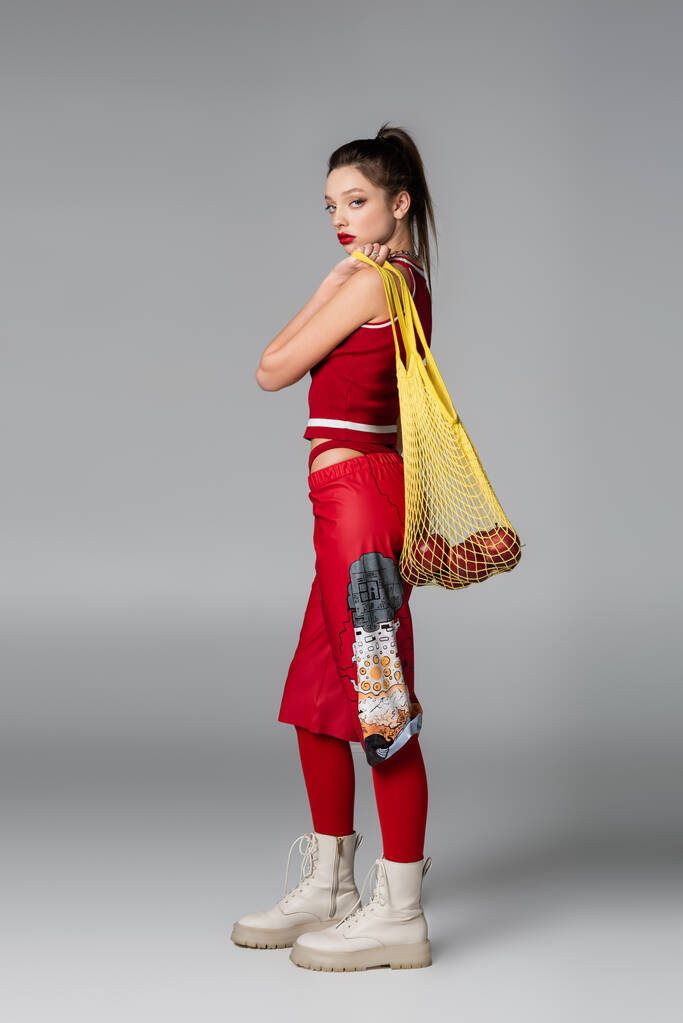 повна довжина модної молодої моделі в червоному спортивному вбранні, що тримає сумку з яблуками на сірому
 - Фото, зображення
