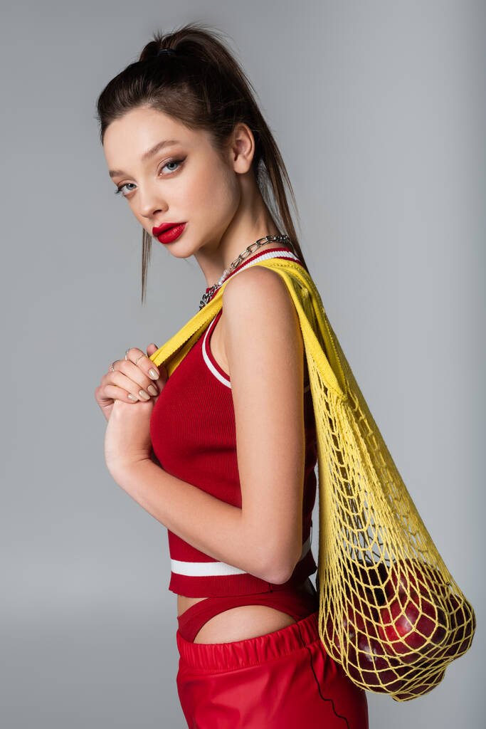 young stylish woman in red sportive outfit holding yellow string bag with apples isolated on grey - Photo, Image