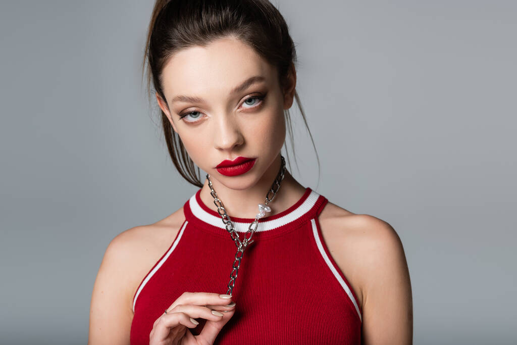 mujer joven y elegante con labios rojos tocando cadena collar aislado en gris - Foto, Imagen