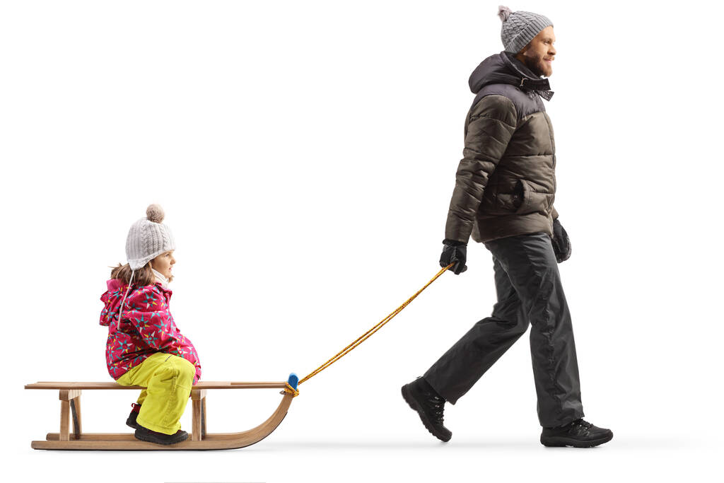 Full length profile shot of a man in winter clothes walking and pulling a girl on a sled isolated on white background - Photo, Image