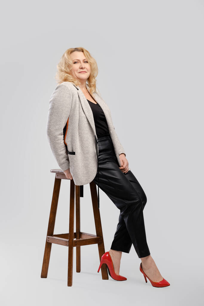 Portrait de femme âgée en pantalon et blazer en cuir assis de profil sur fond de studio gris - Photo, image