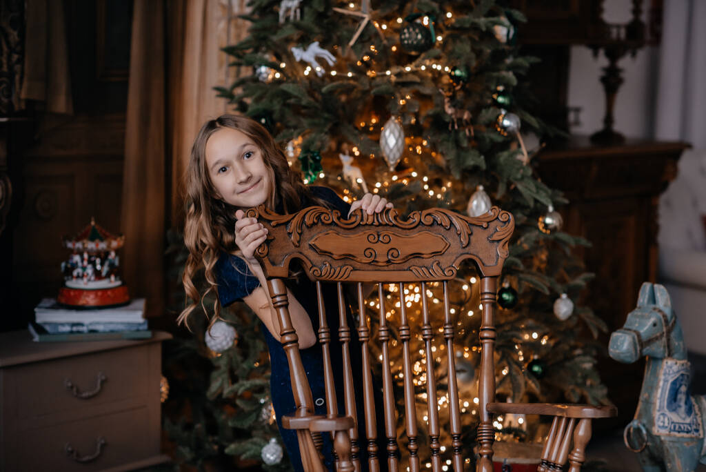 Leuk klein meisje in een blauwe jurk poseert bij de kerstboom. Een kind op een nieuwjaarslocatie. Fotostudio met nieuwjaarsdecoratie. Het kind is in beweging. Selectieve focus, defocus. - Foto, afbeelding