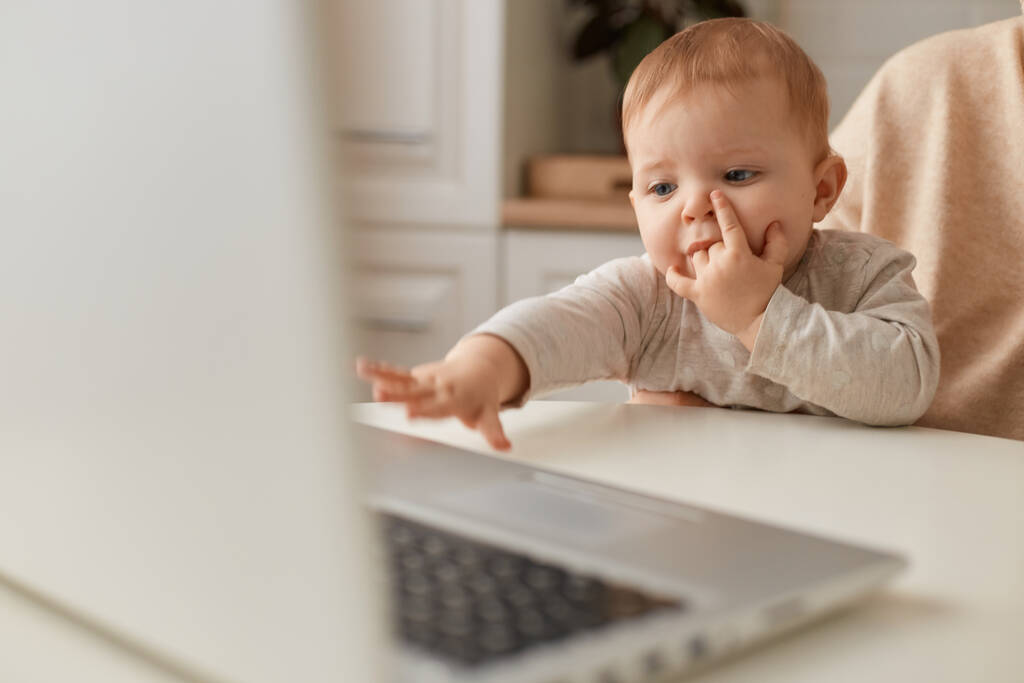 Nahaufnahme Porträt eines Babys, das auf gesichtslosen Mütterknien sitzt und seine Hand zu Laptop, Kindern und moderner Technik ausstreckt. - Foto, Bild