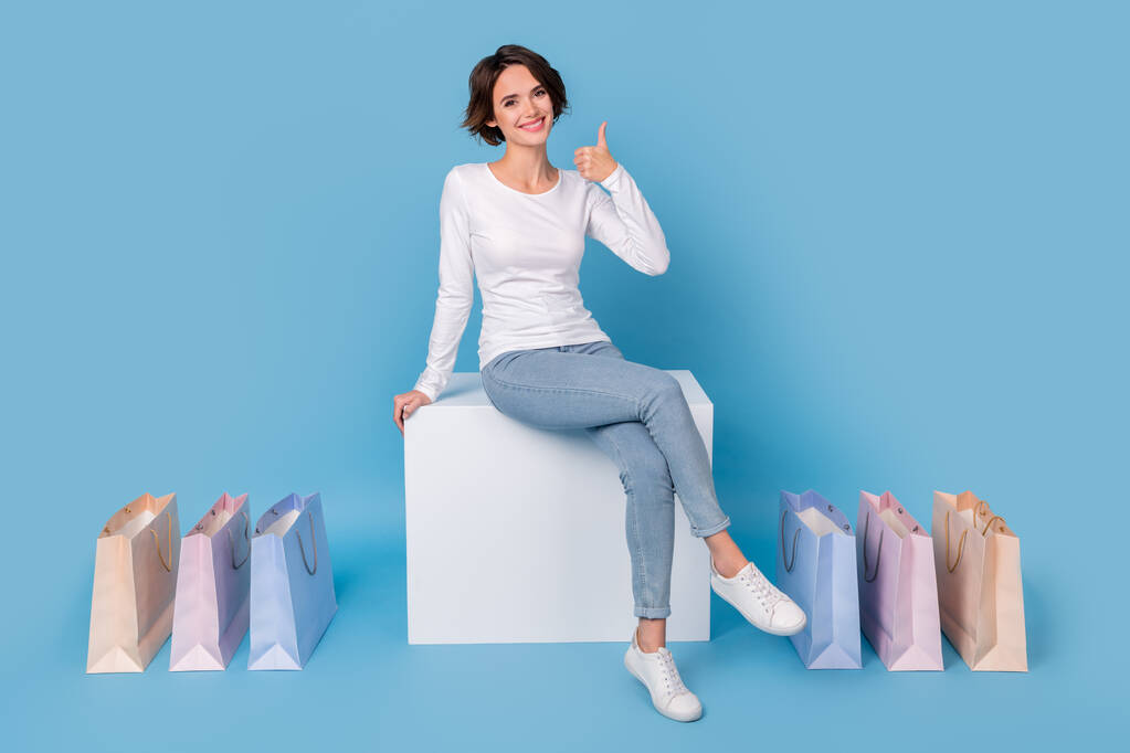 Фото очаровательной сладкой женщины носить белую рубашку сидя белый подиум стул палец вверх изолированный синий цвет фона. - Фото, изображение