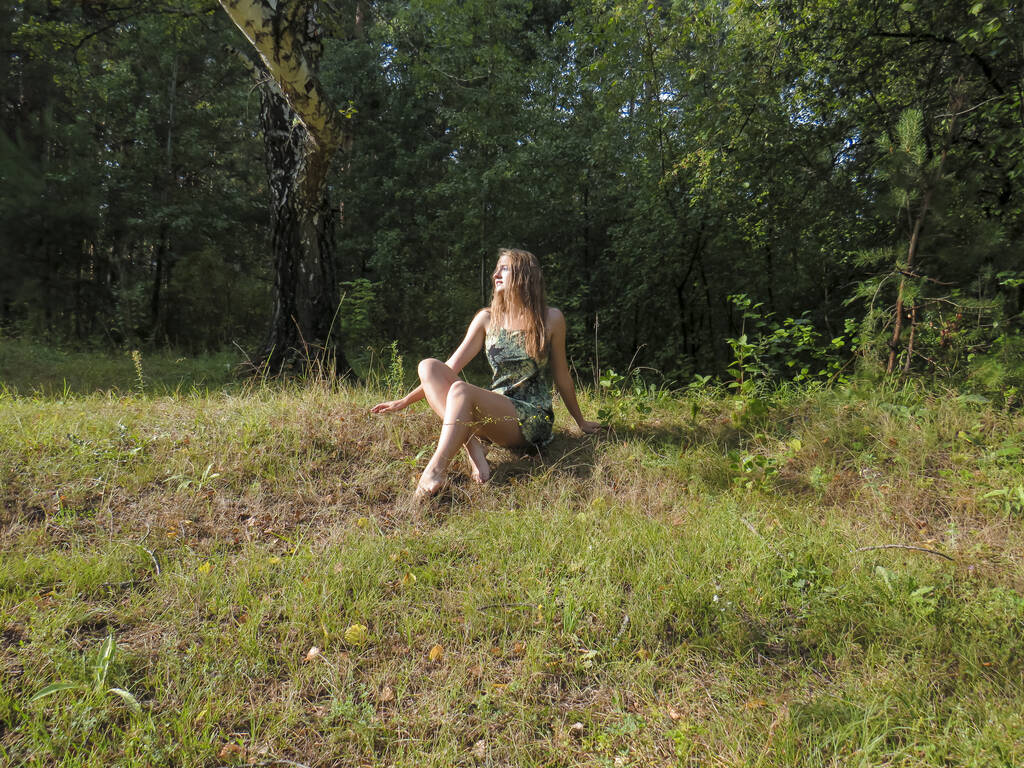 Eine junge rothaarige schöne Frau mit schlanker Figur in einem engen kurzen Kleid mit nackten Schultern ruht an einem warmen, sonnigen Sommertag in den Sonnenstrahlen eines Nadelwaldes auf einem gelben Teppich aus umgefallenen Kiefernnadeln. - Foto, Bild