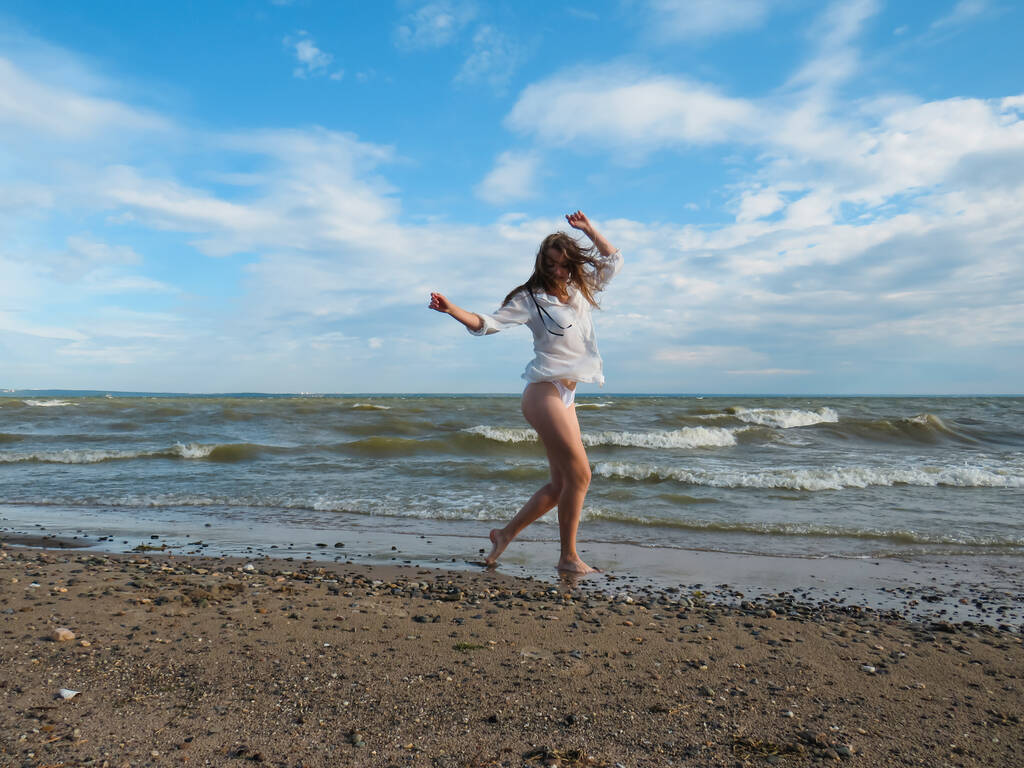 Молодая блондинка с растрепанными волосами в белой рубашке и бикини с тонкими загорелыми ногами на пляже на мокром песке на фоне морских волн и голубого облачного неба в жаркий ветреный летний день, концепция активного образа жизни - Фото, изображение