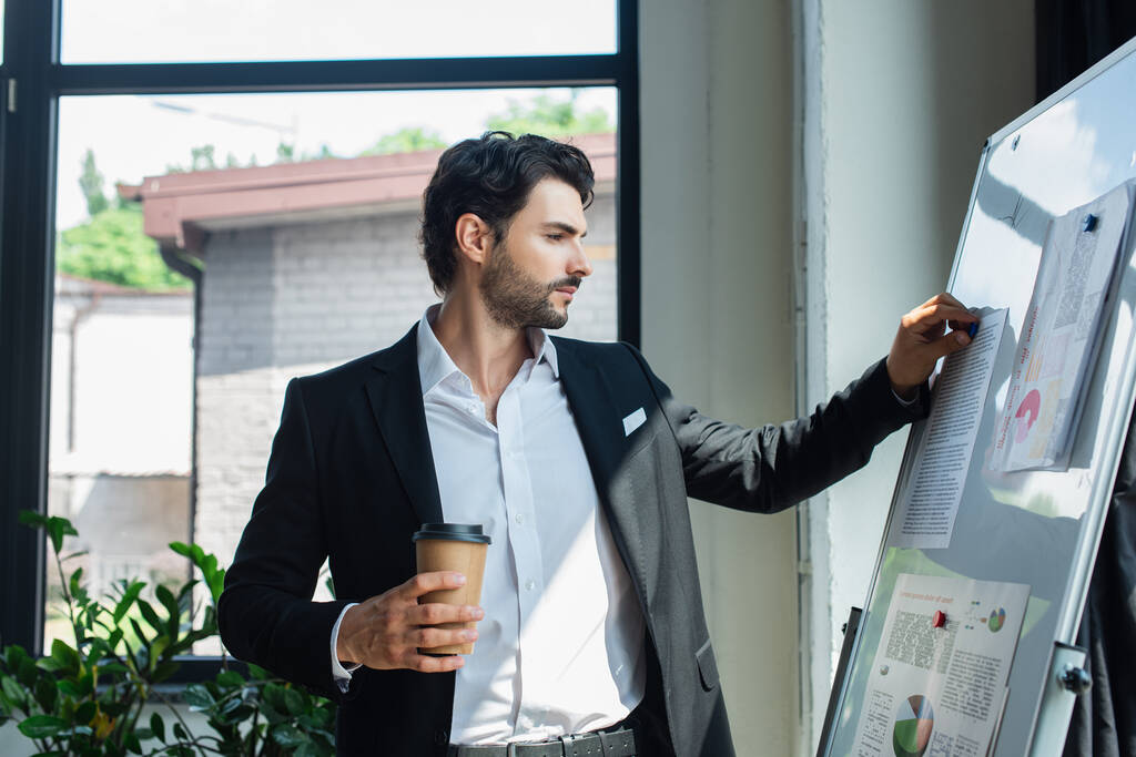 hombre de negocios en blazer negro sosteniendo café para ir cerca de rotafolio con infografías en la oficina - Foto, imagen