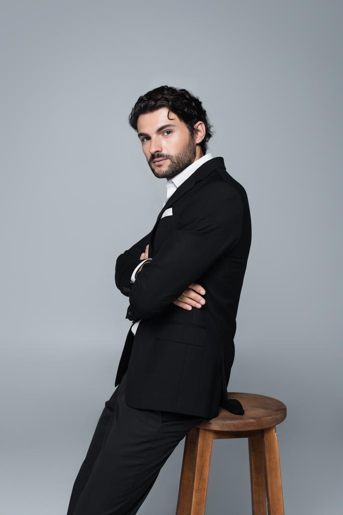 stylový muž v černém obleku stojí poblíž vysoké stoličky se zkříženýma rukama a dívá se na kameru izolovanou na šedi - Fotografie, Obrázek