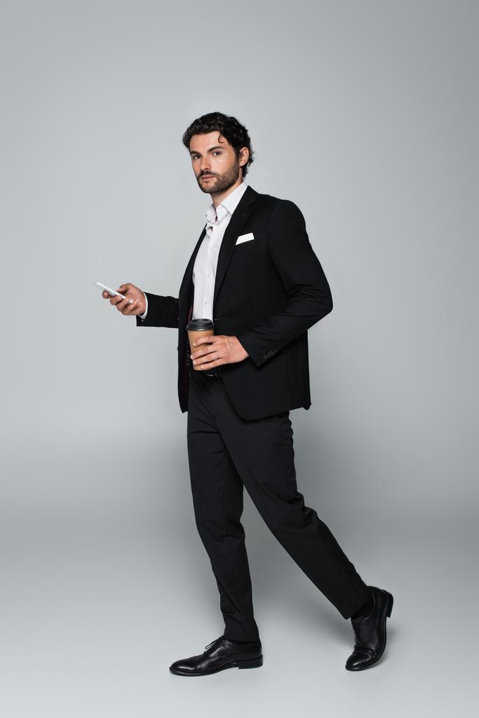 Вид бизнесмена в черном костюме в полный рост, стоящего со смартфоном и бумажной чашкой на сером - Фото, изображение