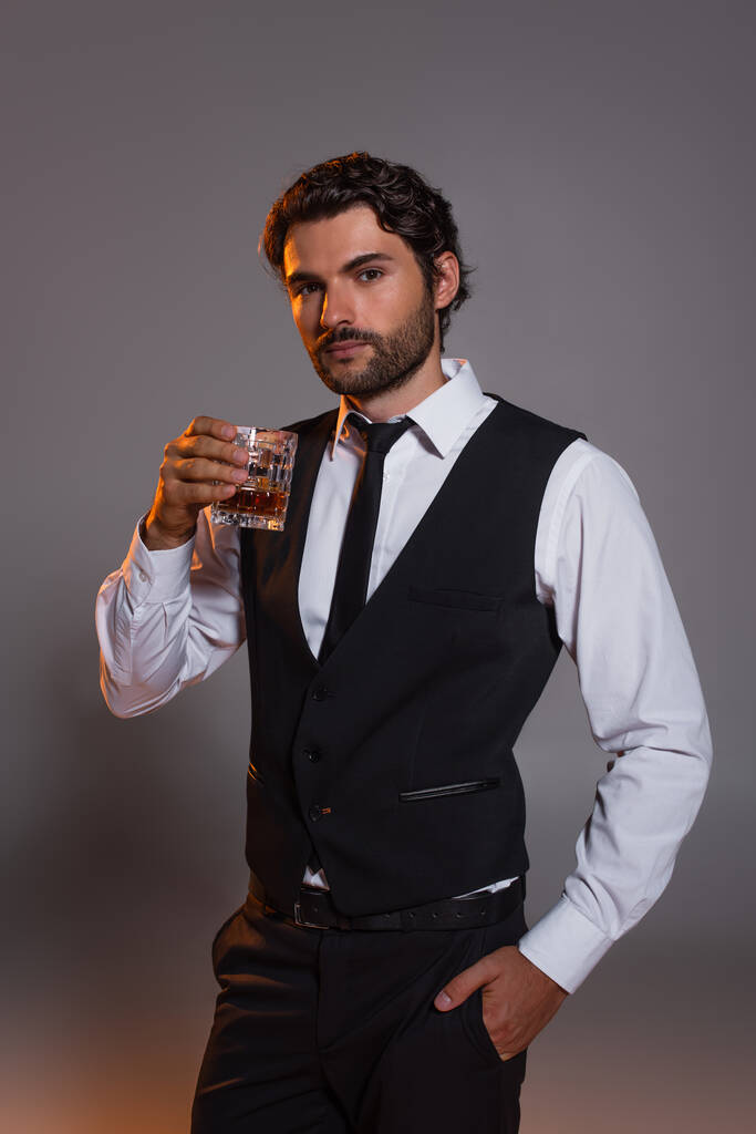 elegante morena hombre con vaso de whisky de pie con la mano en el bolsillo sobre fondo gris - Foto, Imagen