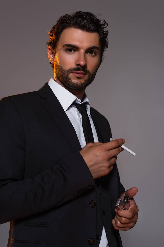 tyylikäs mies mustassa puvussa katselee kameraa pitäen savuketta ja kevyempää eristettynä harmaaseen - Valokuva, kuva