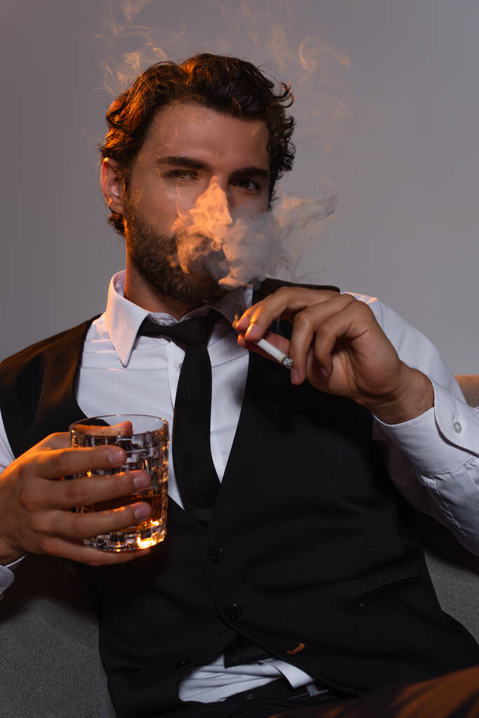 stijlvolle man met glas whisky rooksigaret en kijkend naar camera op grijze achtergrond - Foto, afbeelding