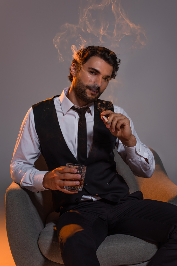 elegante brunette man met glas whisky zitten in fauteuil en roken op grijze achtergrond - Foto, afbeelding