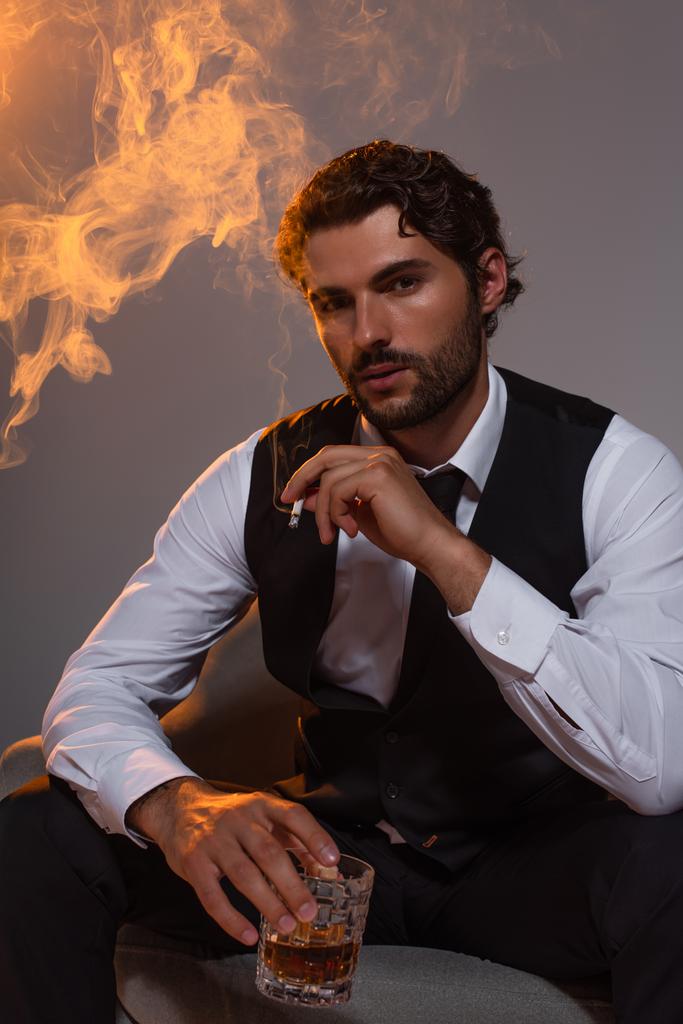 homem elegante sentado em poltrona com copo de uísque e cigarro em fundo cinza com fumaça e luz - Foto, Imagem