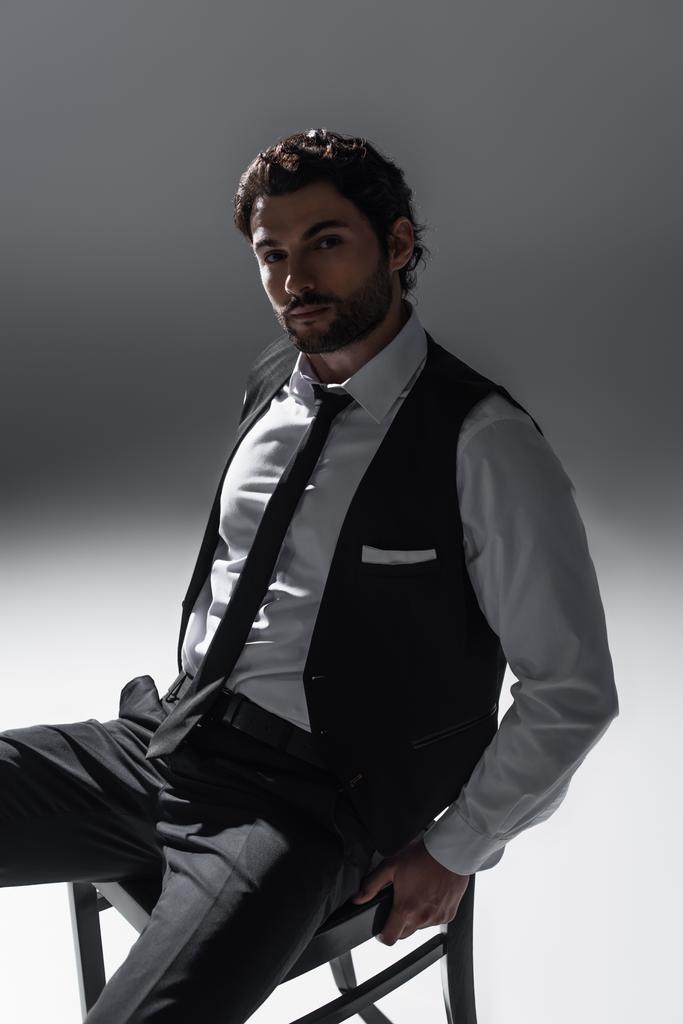 brunetta uomo in elegante abbigliamento formale guardando la fotocamera mentre seduto sul grigio - Foto, immagini