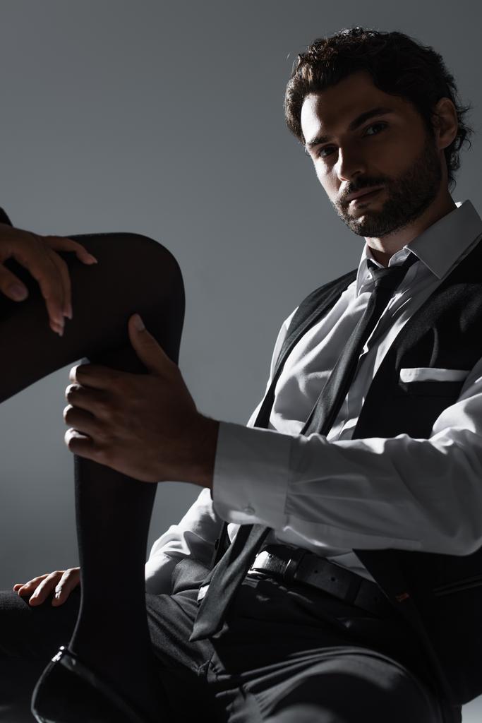 brunette élégant homme toucher jambe de femme sexy tout en regardant caméra isolé sur gris - Photo, image