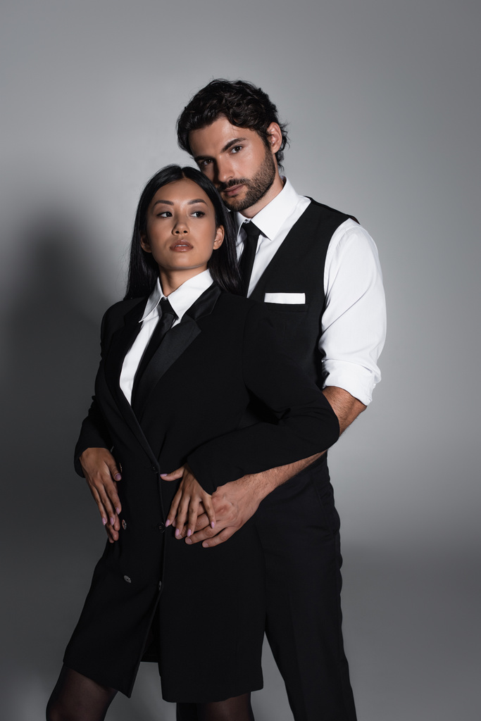 brünetter Mann blickt in die Kamera, während er asiatische Frau in schwarzem Blazer-Kleid auf grauem Hintergrund umarmt - Foto, Bild