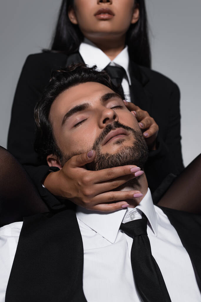 hombre con los ojos cerrados cerca de la mujer sexy seduciéndolo sobre fondo borroso aislado en gris - Foto, Imagen