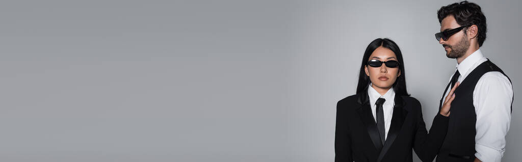donna asiatica in occhiali da sole neri e abbigliamento formale guardando la fotocamera vicino all'uomo alla moda isolato sul grigio, banner - Foto, immagini