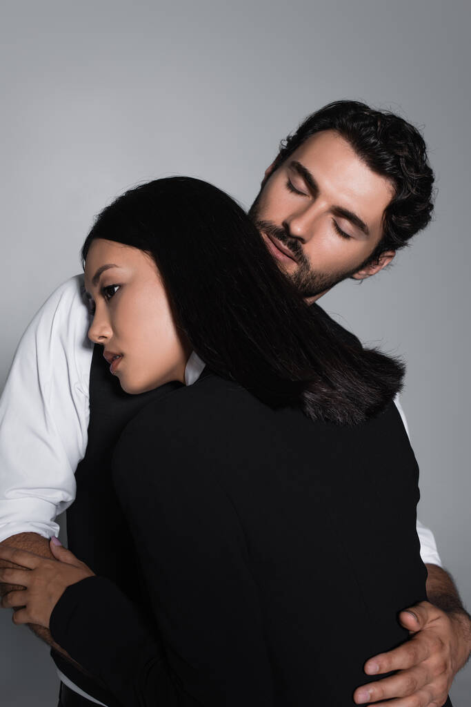 Mann mit geschlossenen Augen umarmt sinnliche asiatische Frau isoliert auf grau - Foto, Bild