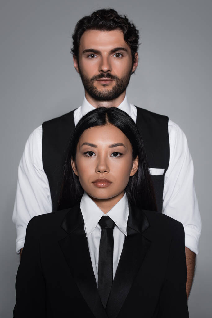 jong aziatische vrouw en brunette man in formele slijtage kijken naar camera geïsoleerd op grijs - Foto, afbeelding