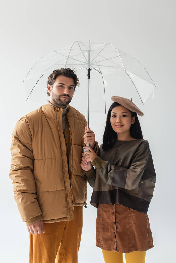 Mann in beigefarbener Pufferjacke und Asiatin im Lederpullover schauen vereinzelt in die Kamera unter Regenschirm auf Grau - Foto, Bild