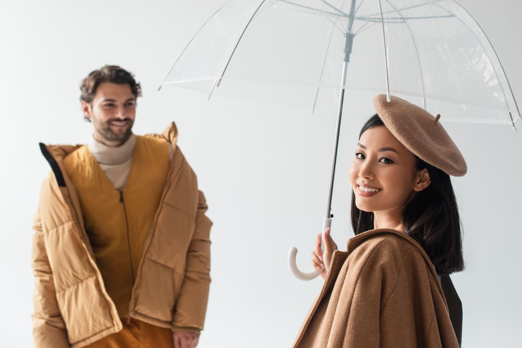 Kameraya gülümseyen şeffaf şemsiyeli Asyalı kadın gri üzerine izole edilmiş bulanık adamın yanında. - Fotoğraf, Görsel