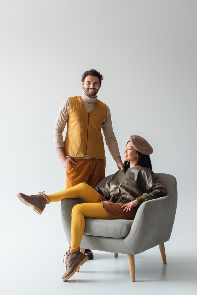 happy and stylish man standing with hand in pocket near asian woman in armchair on grey - Photo, Image