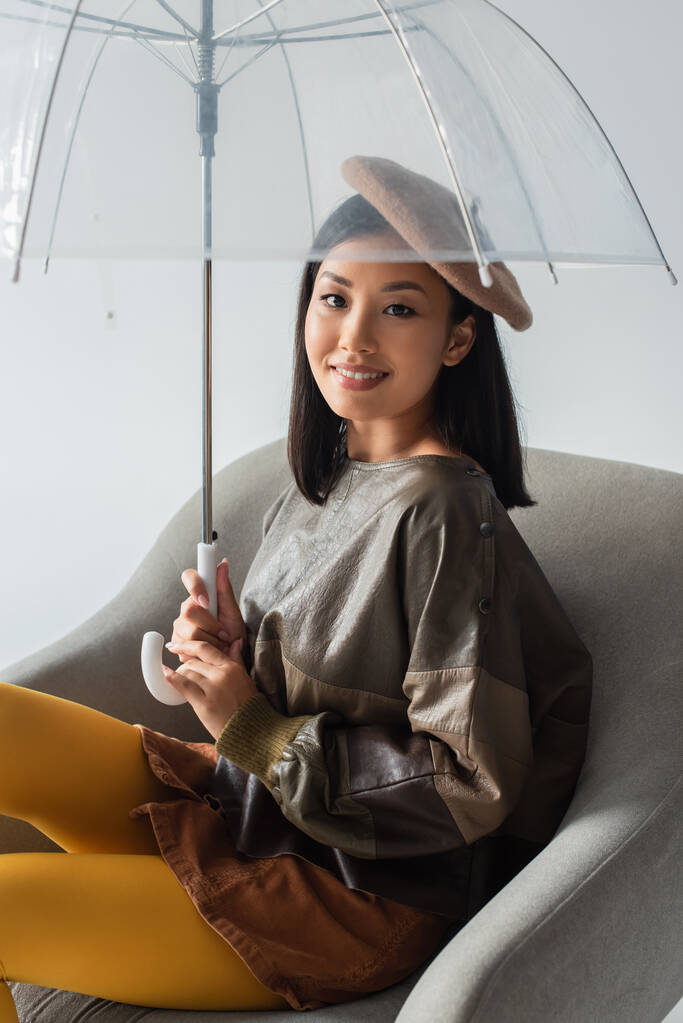 stylish asian woman sitting in armchair under umbrella and smiling at camera isolated on grey - Photo, Image