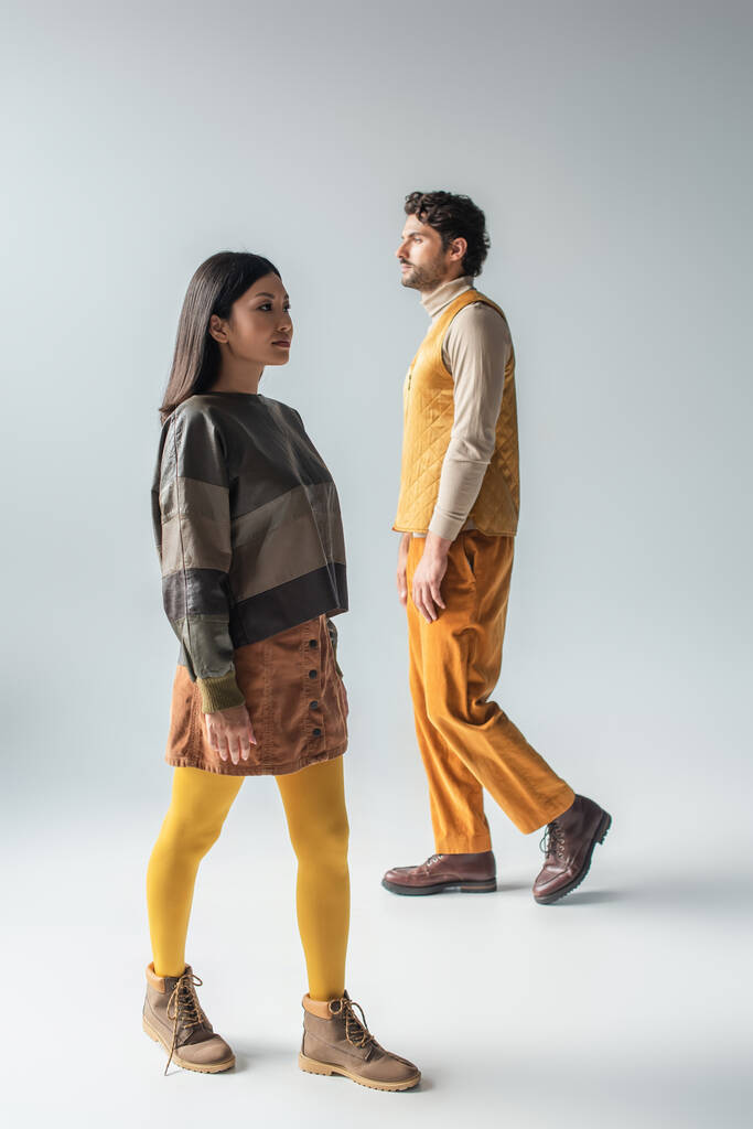 volledige weergave van aziatische vrouw en brunette man in stijlvolle kleding lopen op grijs - Foto, afbeelding